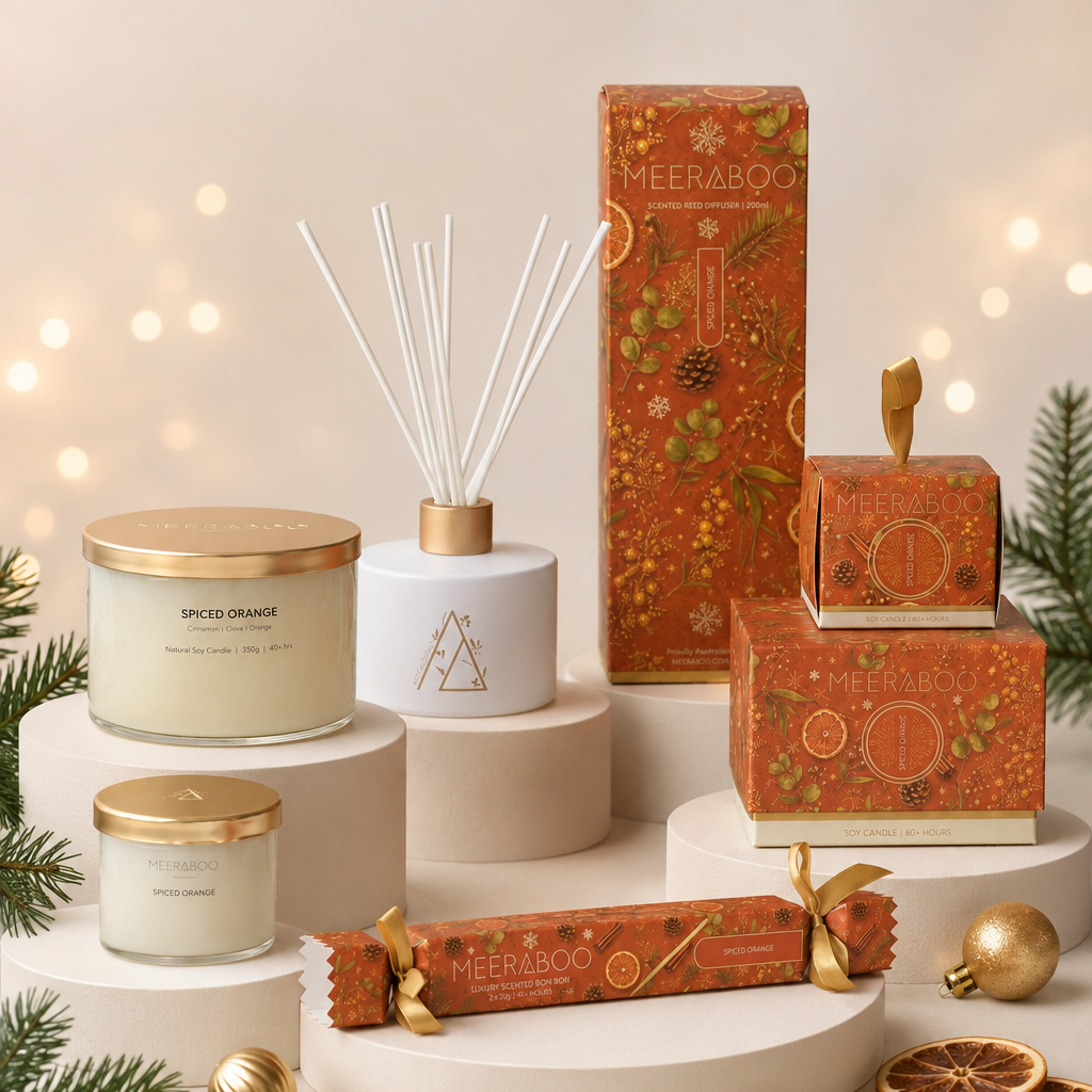 A coordinated holiday gift set from Meeraboo, including a Spiced Orange scented candle, a reed diffuser, two smaller candles, a tall decorative box, a wrapped ornament, and a wrapped gift, arranged on white pedestals with pine branches and warm bokeh lights in the background.